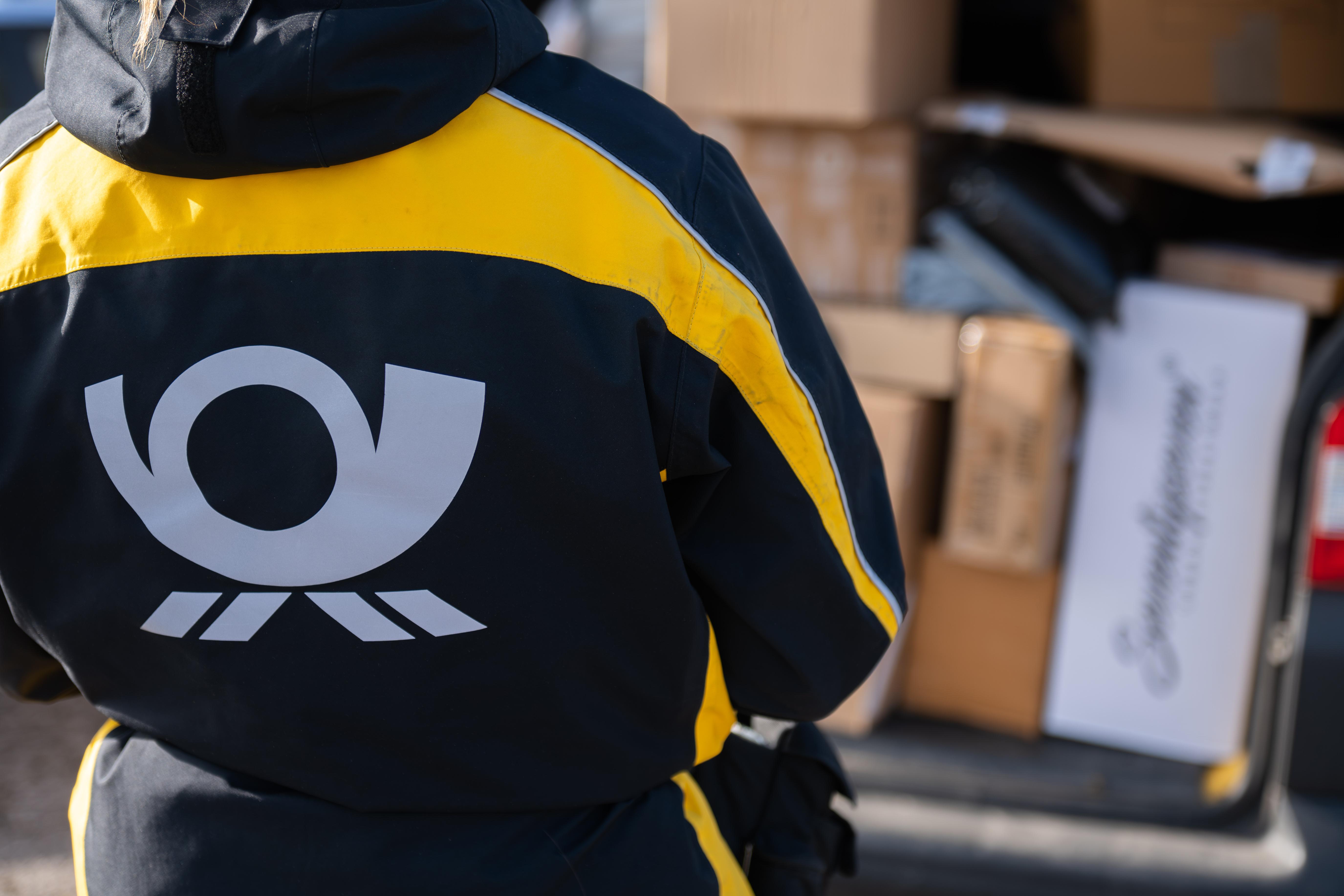 Eine Postbotin trägt eine Jacke mit dem Logo der Deutschen Post und hält dabei ein Paket vor einem Zustellfahrzeug in der Hand.