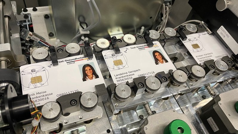In einer Maschine sind Rohlinge von Chipkarten in der Herstellung zu sehen. Darauf zu sehen ist das Bild einer Frau und Aufdrücke wie "Test-Gesundheitskasse", "Landelina Gerspach", Krankenkasse und Zahlenkombinationen.