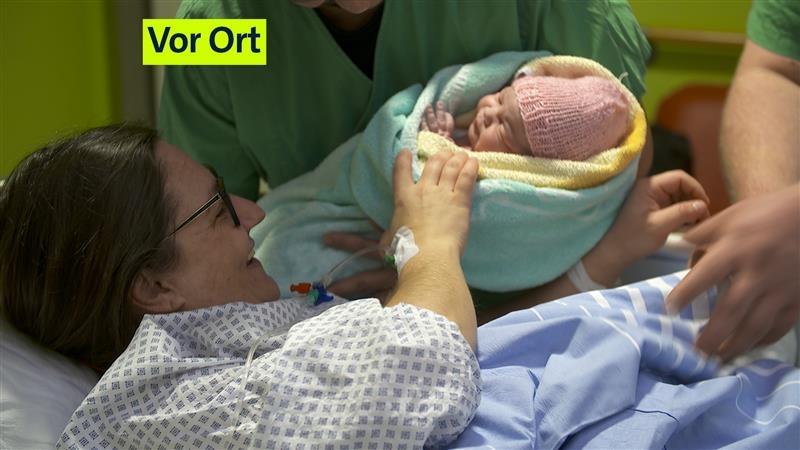 Eine Mutter liegt lächelnd in einem Klinikbett, während sie ihr kleines Baby, das in ein Handtuch eingewickelt ist, übergeben bekommt. 