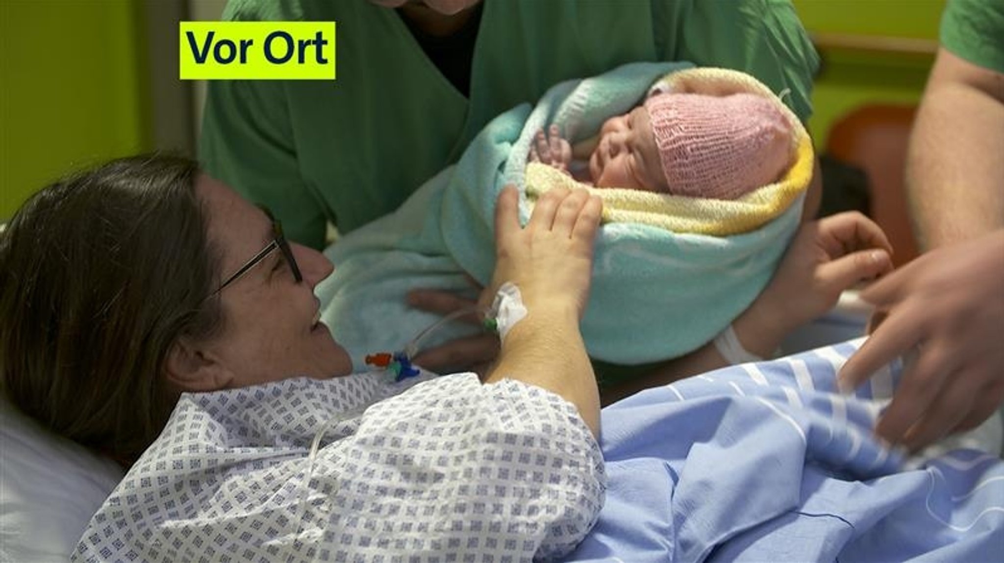 Eine Mutter liegt lächelnd in einem Klinikbett, während sie ihr kleines Baby, das in ein Handtuch eingewickelt ist, übergeben bekommt. 