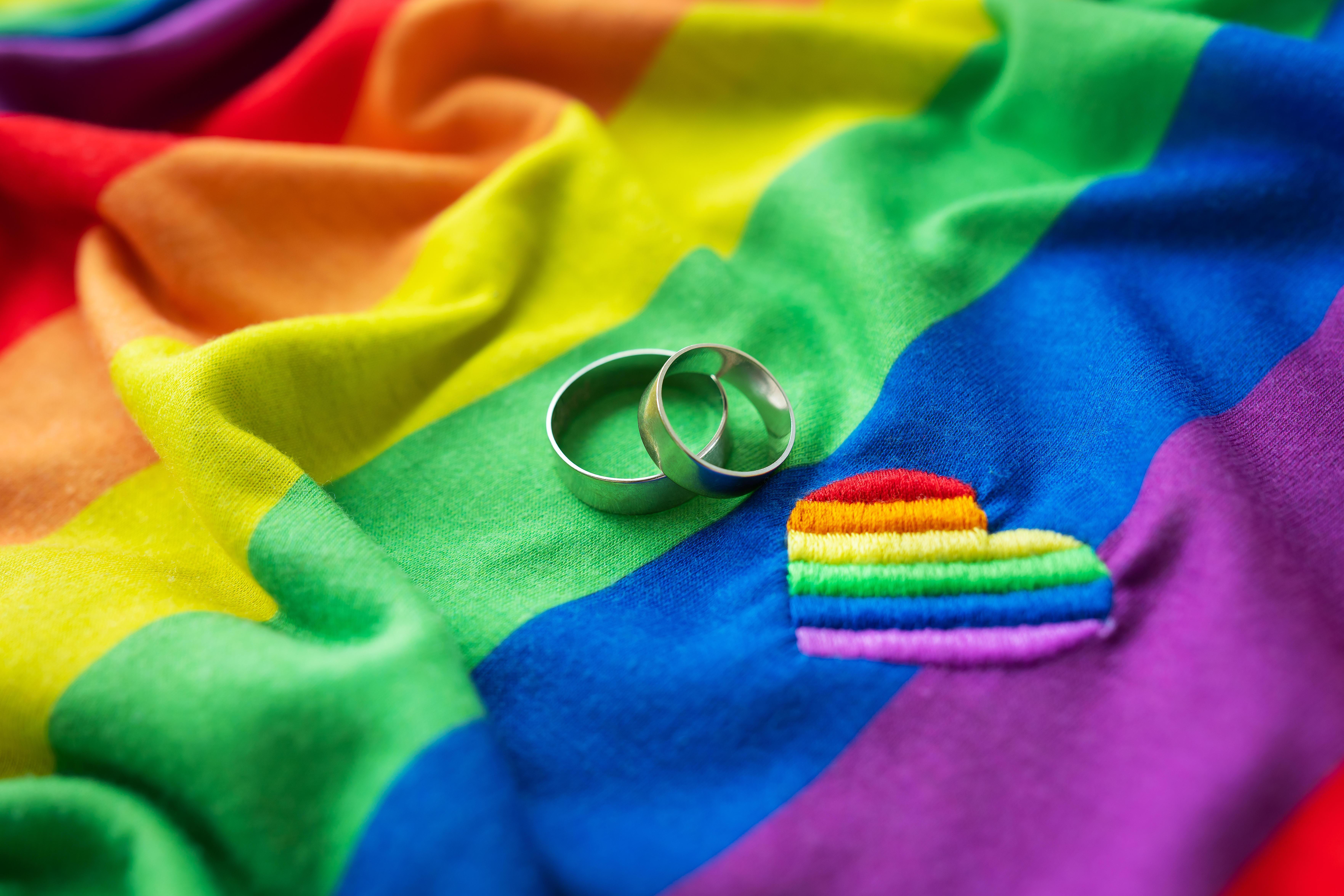 (Symbolbild) Zwei Eheringe auf einer LGBT-Regenbogenflagge, daneben ein aufgesticktes Herz.