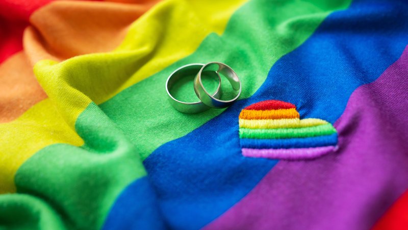 (Symbolbild) Zwei Eheringe auf einer LGBT-Regenbogenflagge, daneben ein aufgesticktes Herz. | Bild: picture alliance / Zoonar | Sergiy Artsaba (Symbolbild) Zwei Eheringe auf einer LGBT-Regenbogenflagge, daneben ein aufgesticktes Herz.
