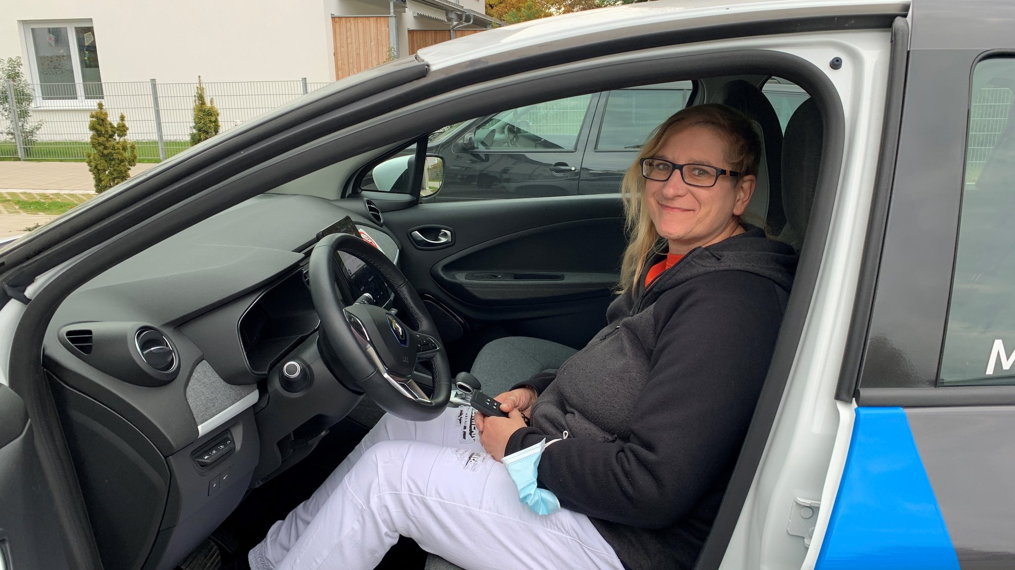 Eine Frau sitzt in einem Auto auf der Fahrerseite und lächelt in die Kamera. 