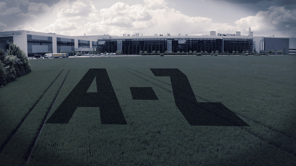 Ein landwirtschaftliches Feld, auf dem "A-Z" projiziert ist, im Hintergrund: Audi-Gebäude in Ingolstadt.