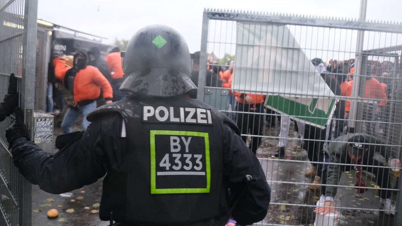 Vor einer Gittertor steht ein Polizeibeamter mit Helm, dahinter vermummte Fußballfans.