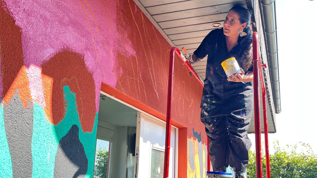  Liliana Martinez mit Farbtopf und Pinsel auf einer Leiter an der Hauswand.
