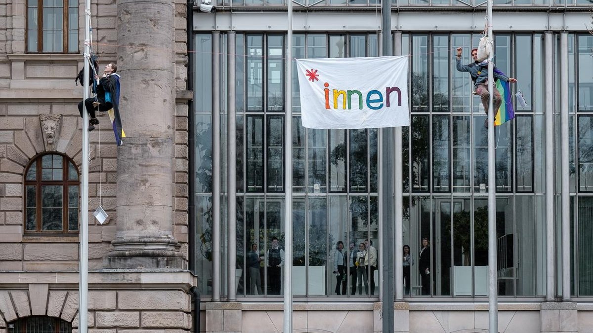 Seit Montag gilt in Bayern das Genderverbot. Doch damit wollen sich zwei junge Menschen in München nicht abfinden. Sie sind am Mittwoch auf die Fahnenmasten vor der Staatskanzlei geklettert - für einen luftigen Protest.