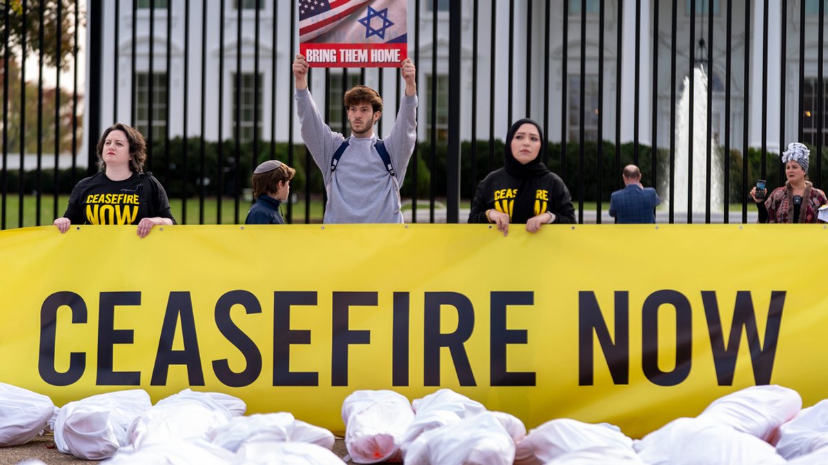 Personen stehen hinter einem Transparent mit "Biden: Ceasefire Now" (Biden: Waffenstillstand jetzt), vor dem imitierte Leichensäcke liegen.