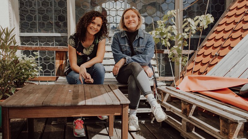 Gründerinnen Nicole Noller aus Asperg und Natalie Stanczak aus Augsburg von der Plattform "Faces of Moms" sitzen auf einer Holzbank auf einer Dachterrasse.