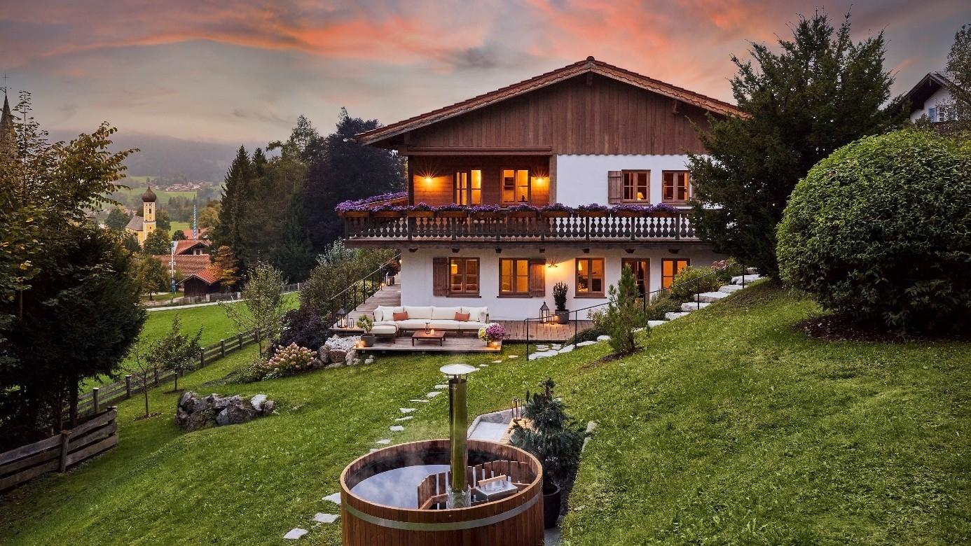 Ein luxuriöses alpenländisches Haus mit Garten an einem Hang vor einer bergigen Landschaft in der Abenddämmerung.