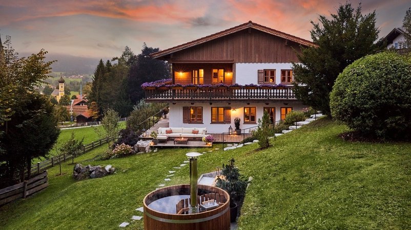 Ein luxuriöses alpenländisches Haus mit Garten an einem Hang vor einer bergigen Landschaft in der Abenddämmerung. | Bild: Omaze Ein luxuriöses alpenländisches Haus mit Garten an einem Hang vor einer bergigen Landschaft in der Abenddämmerung.