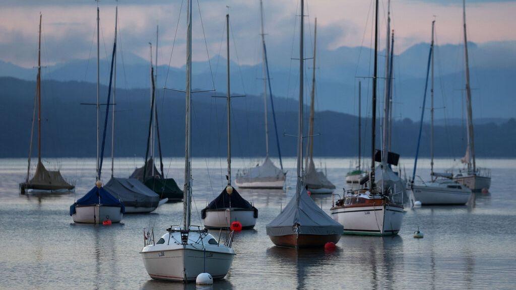 Segelboote liegen im Sonnenaufgang vor dem Panorama der Alpen auf dem Ammersee. 