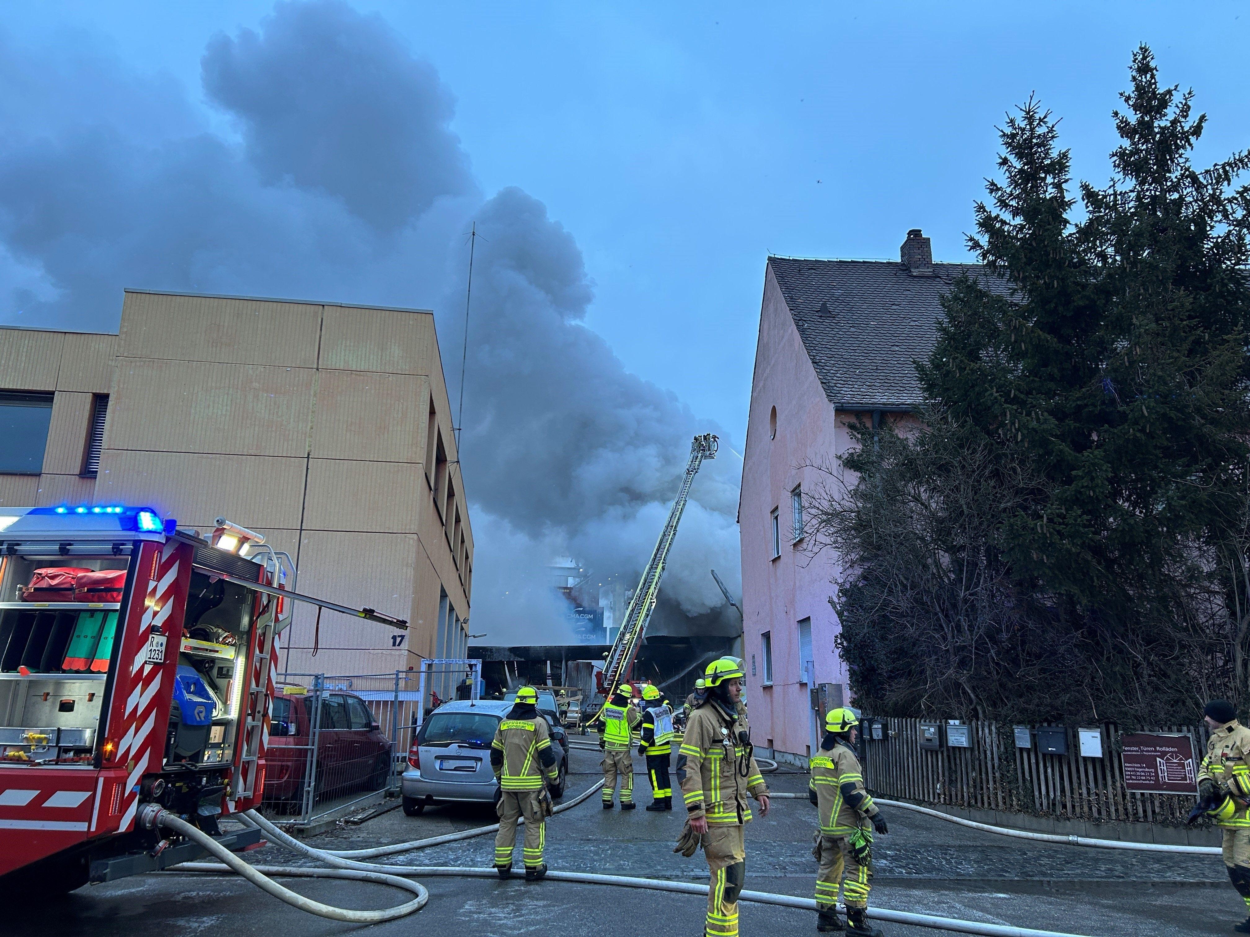 Im Vordergrund Feuerwehrleute in Uniform mit Schläuchen neben einem Feuerwehrwagen. Im Hintergrund eine riesige dicke Rauchsäule hinter zwei Häusern. Eine große Lagerhalle ist am Mittwochmorgen im Regensburger Hafengebiet in Brand geraten.