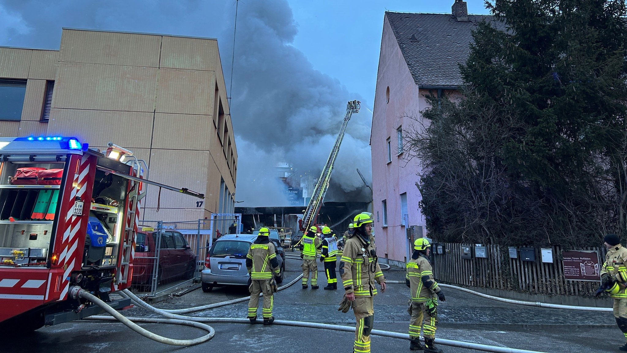 Im Vordergrund Feuerwehrleute in Uniform mit Schläuchen neben einem Feuerwehrwagen. Im Hintergrund eine riesige dicke Rauchsäule hinter zwei Häusern. Eine große Lagerhalle ist am Mittwochmorgen im Regensburger Hafengebiet in Brand geraten.