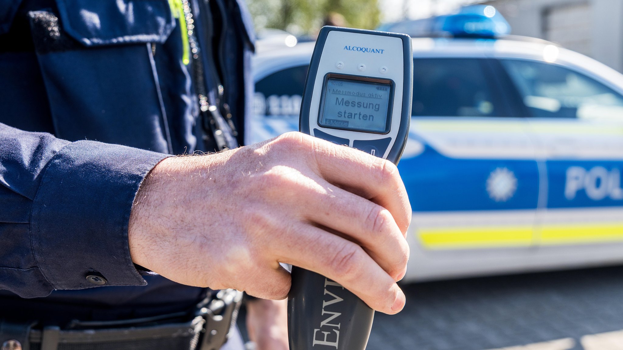 Polizist hält ein Alkoholmessgerät vor einen Streifenwagen 