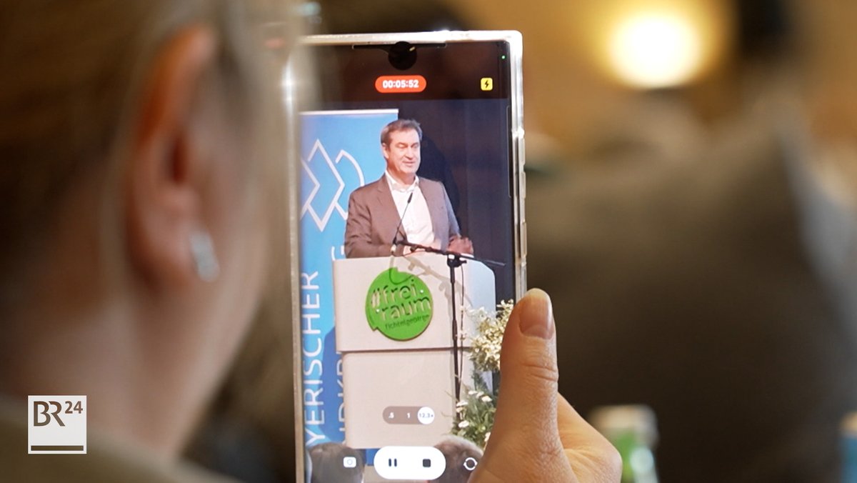 Auf dem Display eines Handys ist Ministerpräsident Markus Söder an einem Rednerpult zu sehen.