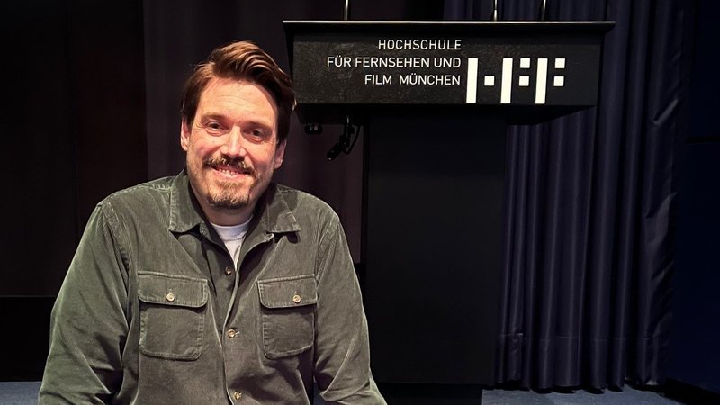 Drehbuchautor Moritz Binder sitzt lächelnd am Rand einer schwarzen Bühne vor einem Rednerpult mit dem Schriftzug "Hochschule für Fernsehen und Film München" | Bild: BR/Markus Aicher Drehbuchautor Moritz Binder sitzt lächelnd am Rand einer schwarzen Bühne vor einem Rednerpult mit dem Schriftzug "Hochschule für Fernsehen und Film München"