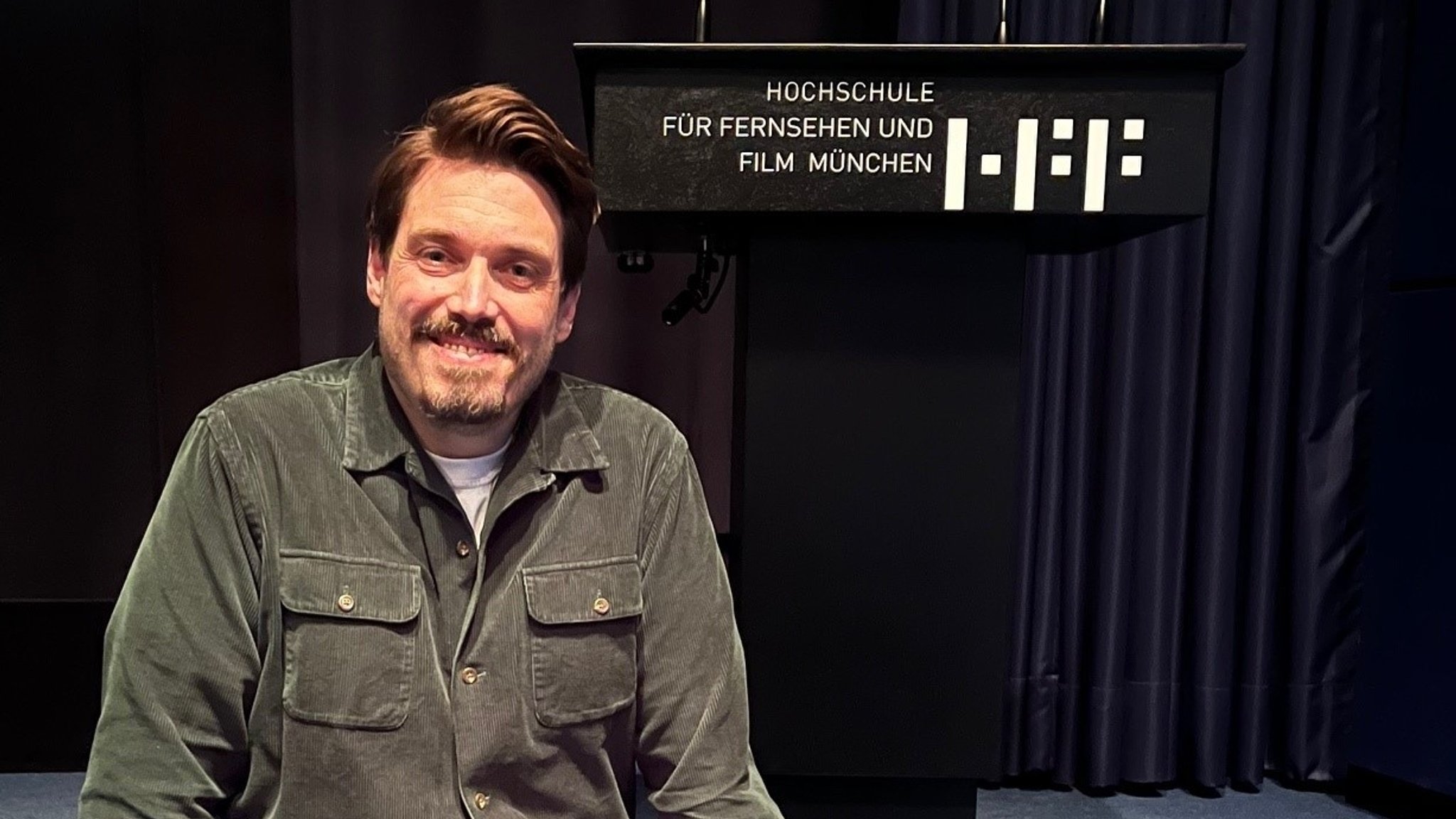 Drehbuchautor Moritz Binder sitzt lächelnd am Rand einer schwarzen Bühne vor einem Rednerpult mit dem Schriftzug "Hochschule für Fernsehen und Film München"