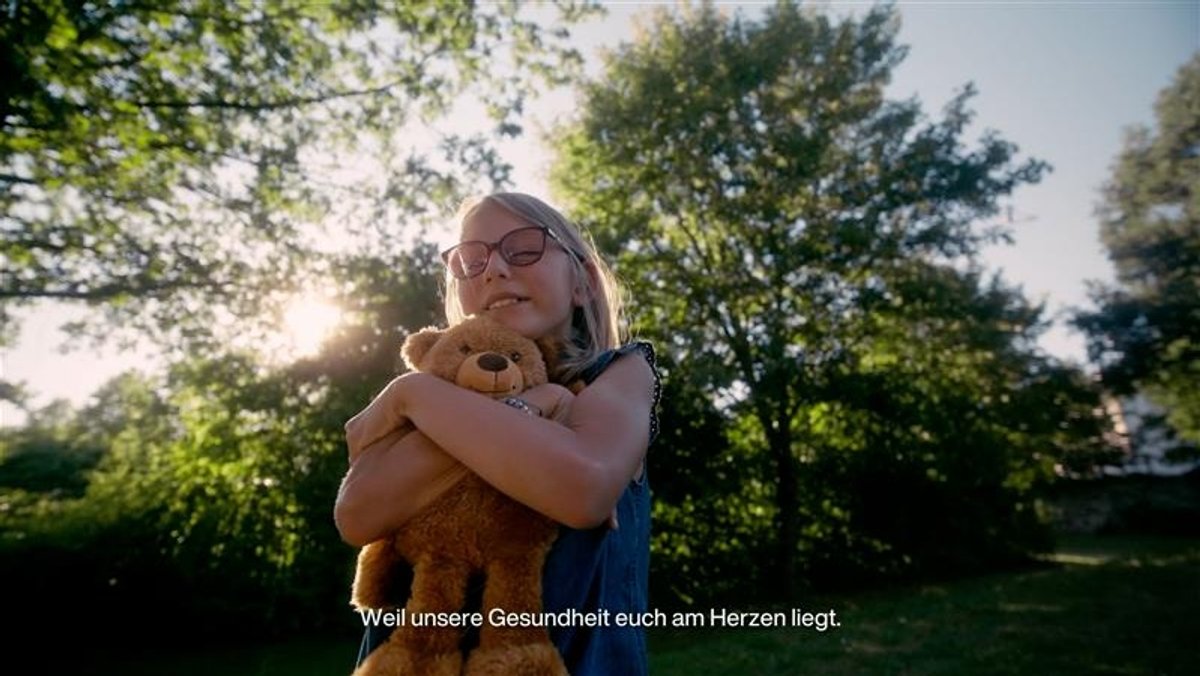 Ausschnitt aus dem Werbespot. Ein Kind vor sommerlicher Baumkulisse umarmt einen Teddybären