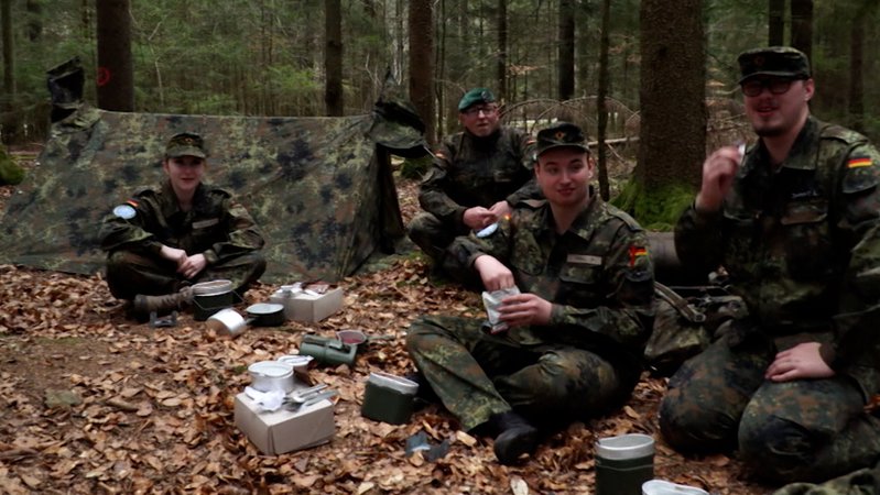 Vier Personen in Bundeswehr-Uniform sitzen auf einem Waldboden, umgeben von Proviant-Ausrüstung der Bundeswehr und einem Zelt in Tarnfarben. | Bild: Bayerischer Rundfunk 2025 Vier Personen in Bundeswehr-Uniform sitzen auf einem Waldboden, umgeben von Proviant-Ausrüstung der Bundeswehr und einem Zelt in Tarnfarben.