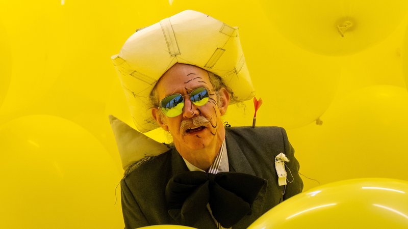 Ein Mann mit verspiegelter Sonnenbrille sitzt in einem Bad aus gelben Luftballons