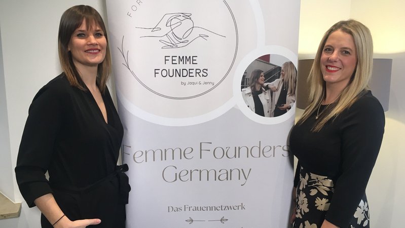 Zwei Frauen stehen grinsend vor einem Plakat der "Femme Founders Germany". 