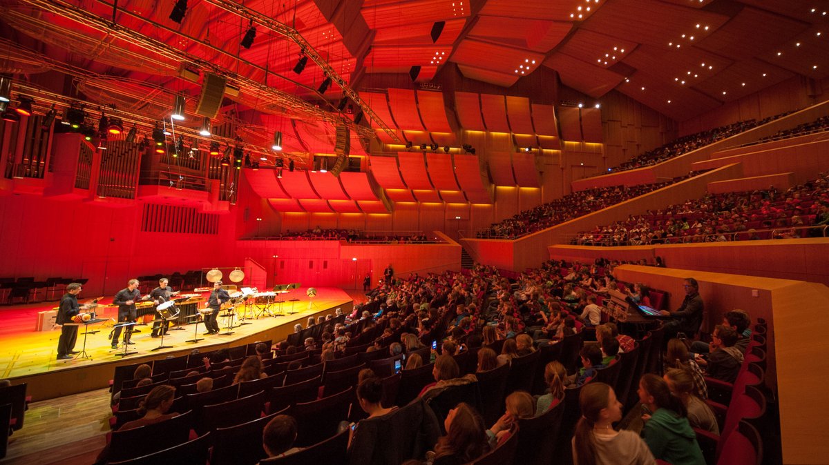 Publikum lauscht Musikern auf der Bühne eines rot ausgeleuchteten Konzertsaals. Laut einer Studie der TU Berlin beeinflusst die Farbe eines Konzertsaals die Wahrnehmung der Musik. Das Auge hört mit, die Farbe bestimmt den Klang der Musik.