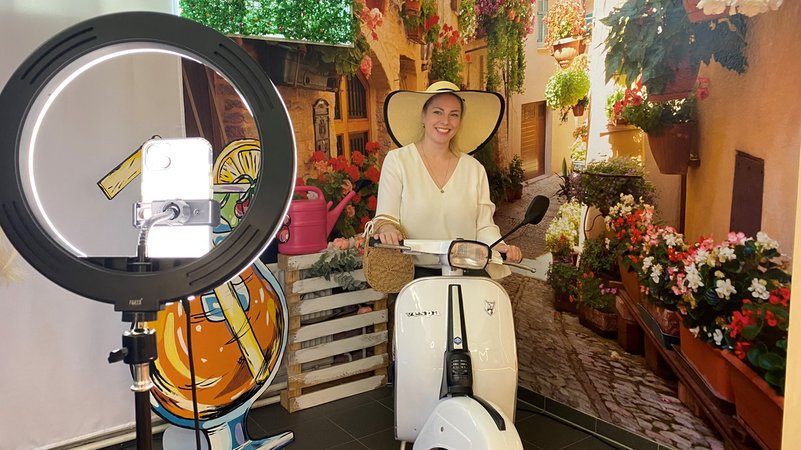 Eine Frau sitzt auf einem Roller vor einer Kulisse. Fotografiert wird sie von einem Mobiltelefon, das in einen Leuchtrahmen gespannt ist. | Bild: BR/Anja Bischof Eine Frau sitzt auf einem Roller vor einer Kulisse. Fotografiert wird sie von einem Mobiltelefon, das in einen Leuchtrahmen gespannt ist.