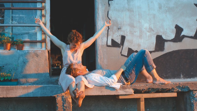 Auf einer Steinbank sitzt ein Paar im Sonnenlicht vor einem offenen Haus. Szene aus "Die Reste meines Lebens" 