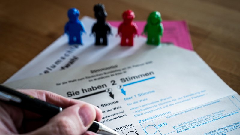 Der Wahl-Stimmzettel mit einer Hand, die einen Stift hält. Am oberen Rand des Zettels stehen vier Spielzeugfiguren in unterschiedlichen Farben. | Bild: picture alliance / Jens Krick Der Wahl-Stimmzettel mit einer Hand, die einen Stift hält. Am oberen Rand des Zettels stehen vier Spielzeugfiguren in unterschiedlichen Farben.
