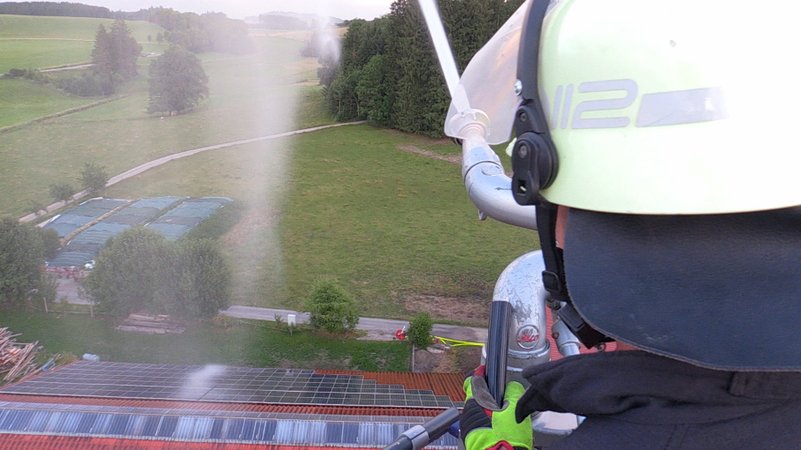 Feuerwehrmann auf einer Drehleiter hoch über einer Scheune mit Photovoltaikanlage. 