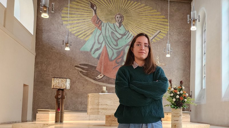 Studentin Lisa Baumeister steht mit verschränkten Armen vor dem Altar. Im Hintergund an der Wand ein Bild von Jesus. | Bild: BR/ Jona Gebhard Studentin Lisa Baumeister steht mit verschränkten Armen vor dem Altar. Im Hintergund an der Wand ein Bild von Jesus.