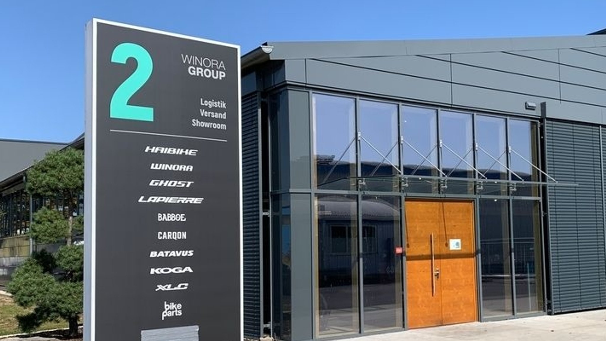 Zu sehen ist ein Gebäude der Winora Group in Sennfeld. Im Vordergrund steht ein großes Hinweisschild mit der Aufschrift „WINORA GROUP“ sowie den Marken Haibike, Winora, Ghost und weiteren Fahrradmarken. Dahinter befindet sich ein modernes, grau verkleidetes Gebäude mit großflächigen Fenstern und einem Eingangsbereich mit Holztür. Auf dem Schild ist „Logistik, Versand, Showroom“ angegeben. Trotz Insolvenz der bayerischen Fahrradhersteller mit ihren bekannten Marken Winora, Haibike und Ghost soll der Betrieb weitergehen. 