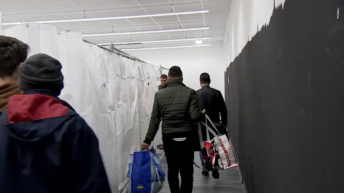 Männer mit Taschen betreten eine zur Notunterkunft umfunktionierte Halle. 