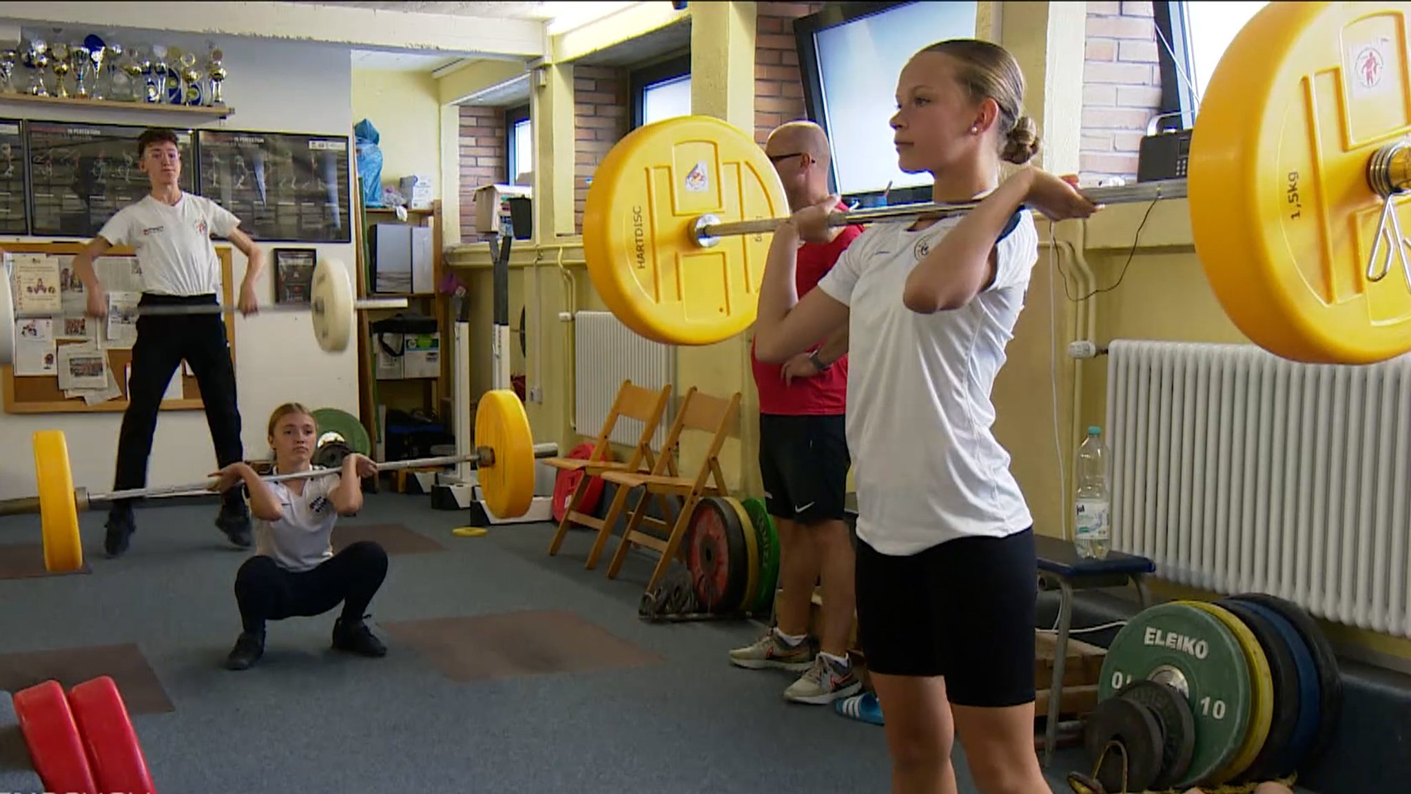 Drei Jugendliche heben in einem Trainingsraum lange Hantelstangen.