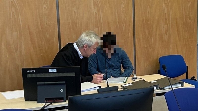 Ein Mann mit gepixeltem Gesicht sitzt auf der Anklagebank neben seinem Anwalt. 