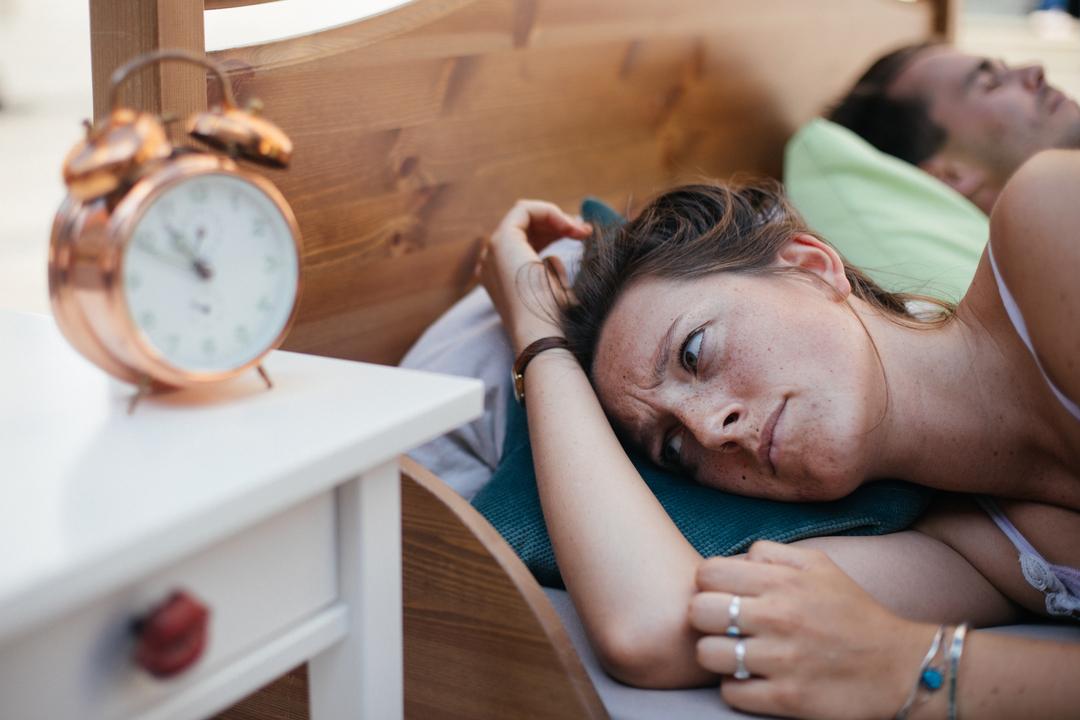 Ein heterosexuelles Paar liegt im Bett. Der Mann schläft, die Frau ist wach und blickt ärgerlich auf den Wecker. Frau 