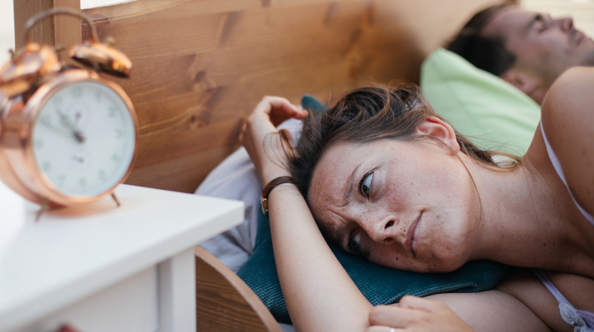 Ein heterosexuelles Paar liegt im Bett. Der Mann schläft, die Frau ist wach und blickt ärgerlich auf den Wecker. Frau 