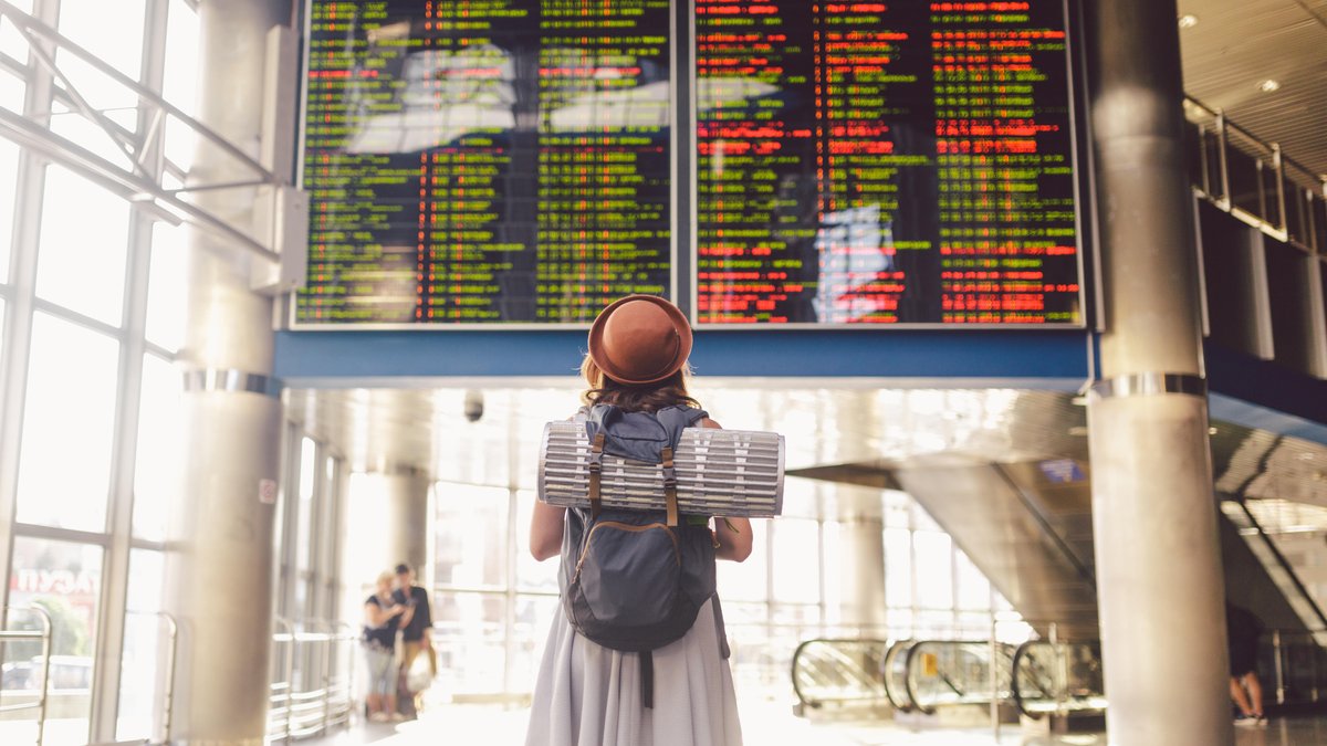 Eine Frau mit Sonnenhut und Rucksack steht in einem Flughafen vor der Abflugtafel.