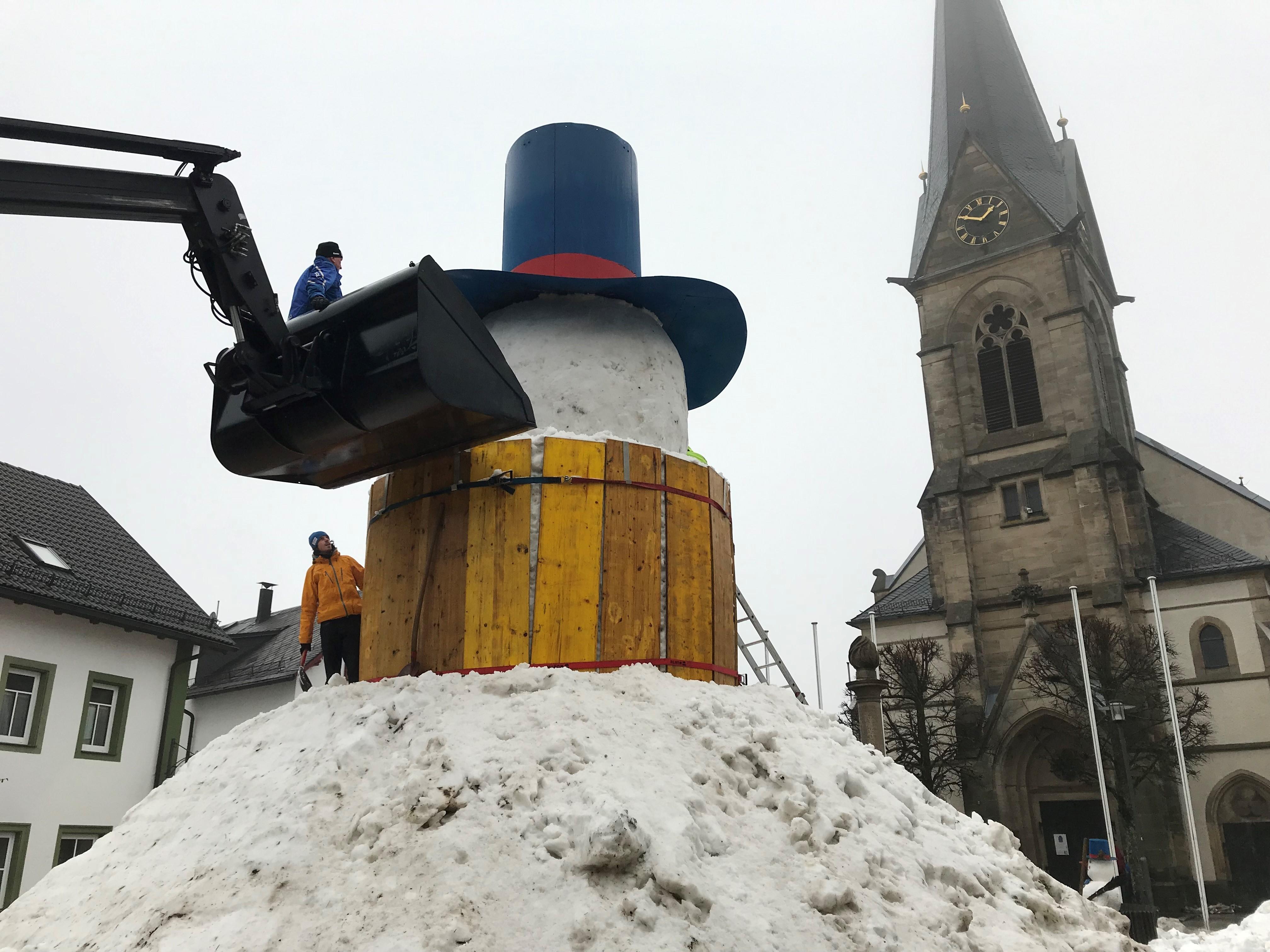 Ein Riesenschneemann noch ohne Gesicht, aber mit Hut, auf dem ein Mann steht und arbeitet, dahinter die Kirche von Bischofsgrün. 