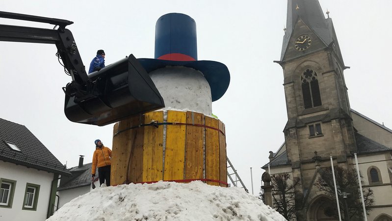 Ein Riesenschneemann noch ohne Gesicht, aber mit Hut, auf dem ein Mann steht und arbeitet, dahinter die Kirche von Bischofsgrün. 
