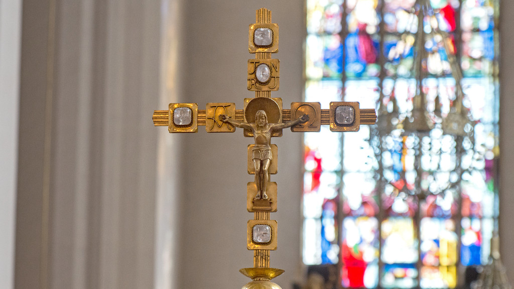 Vortragekreuz aus Messing mit Corpus Christi, eingelassen sind Bergkristalle für die fünf Wunden Christi. Im Hintergrund ein bunt verglastes Kirchenfenster. Aufnahme aus der Münchner Frauenkirche (Symbolbild). 