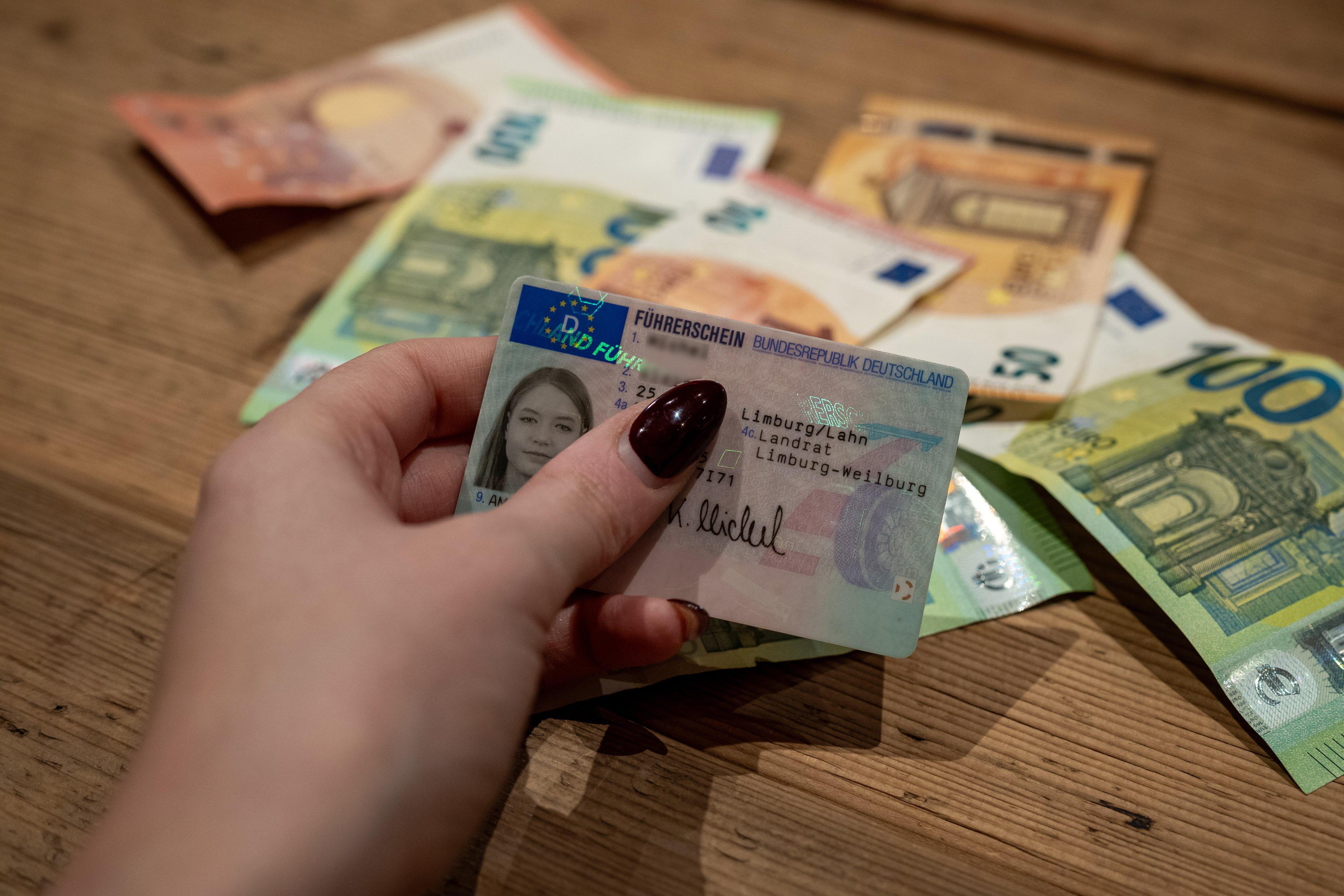 Symbolfoto: Eine junge Frau hält einen Führerschein in der Hand, dahinter liegen Geldscheine.