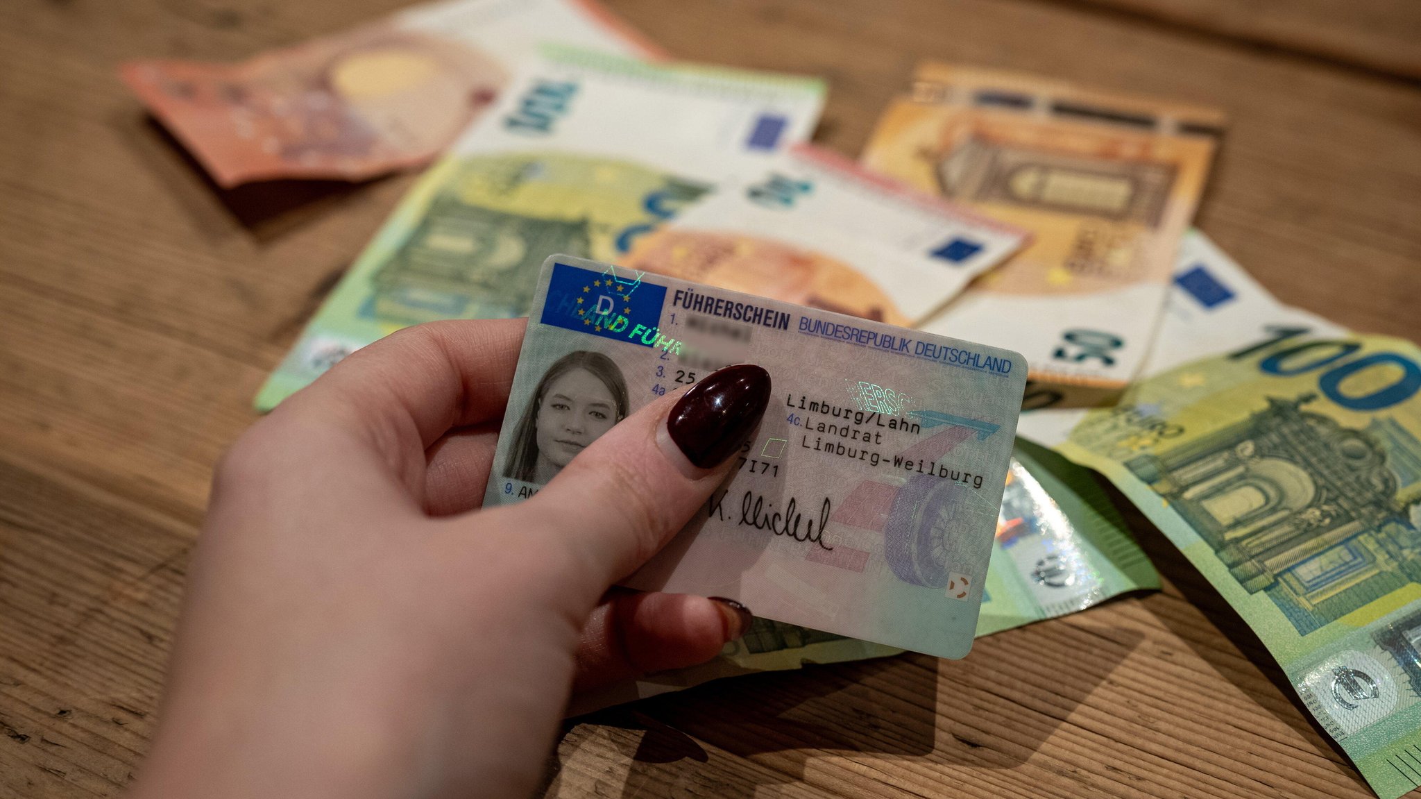 Symbolfoto: Eine junge Frau hält einen Führerschein in der Hand, dahinter liegen Geldscheine.