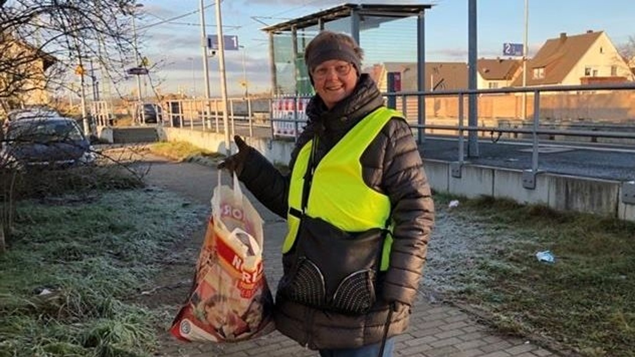 Eine Frau steht vor einem Bahnsteig und hält eine Plastiktüte und eine Müllzange in der Hand. Sie trägt eine gelbe Weste. Im Hintergrund liegt Müll.