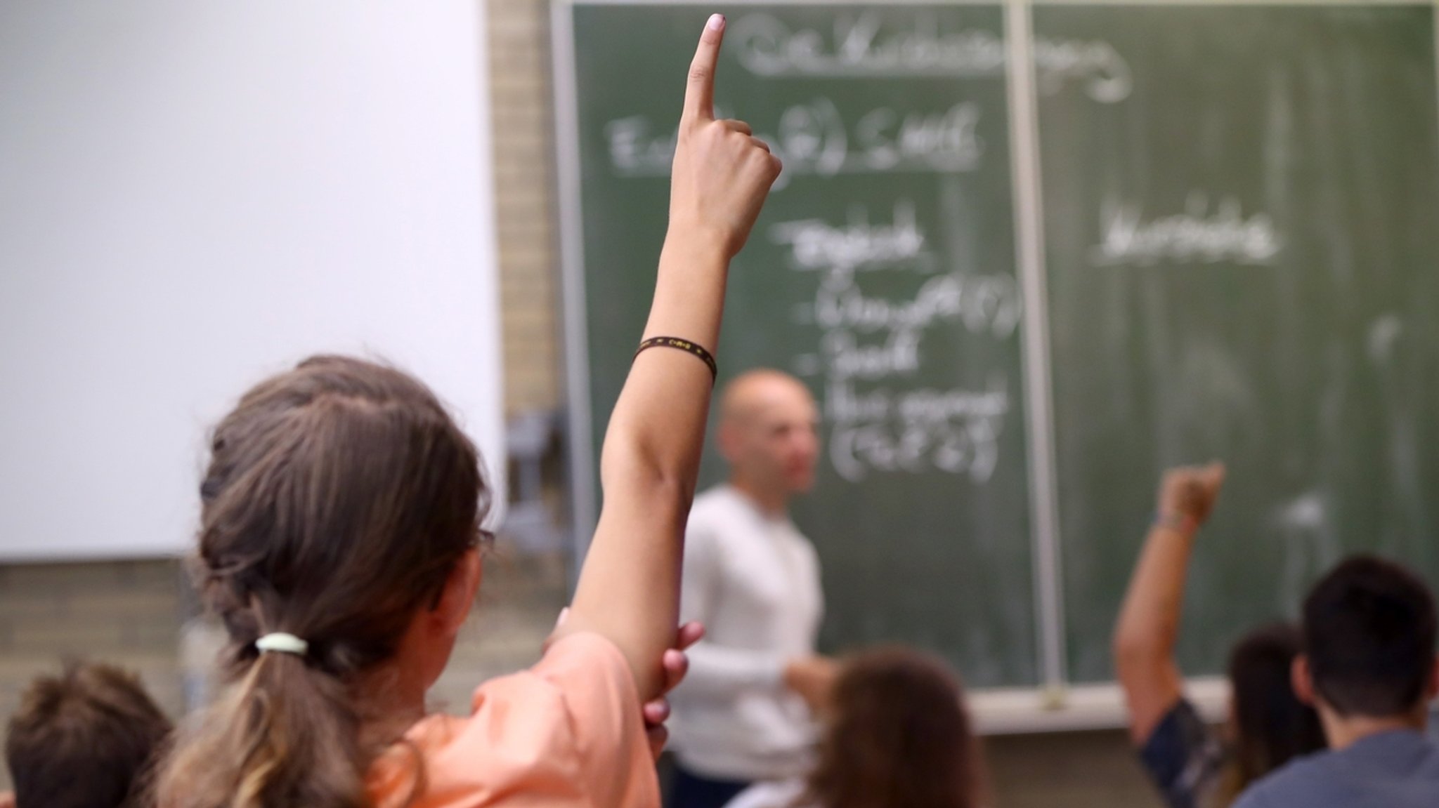 Mädchen von hinten in einer Schulklasse, die den Arm noch oben streckt und sich meldet