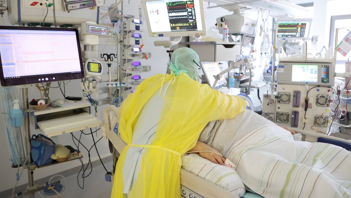 Eine Person liegt im Bett auf einer Intensivstation. Ein Pfleger in Schutzkleidung beugt sich über ihn. Um die beiden stehen viele Geräte unter anderem mit Bildschirmen.