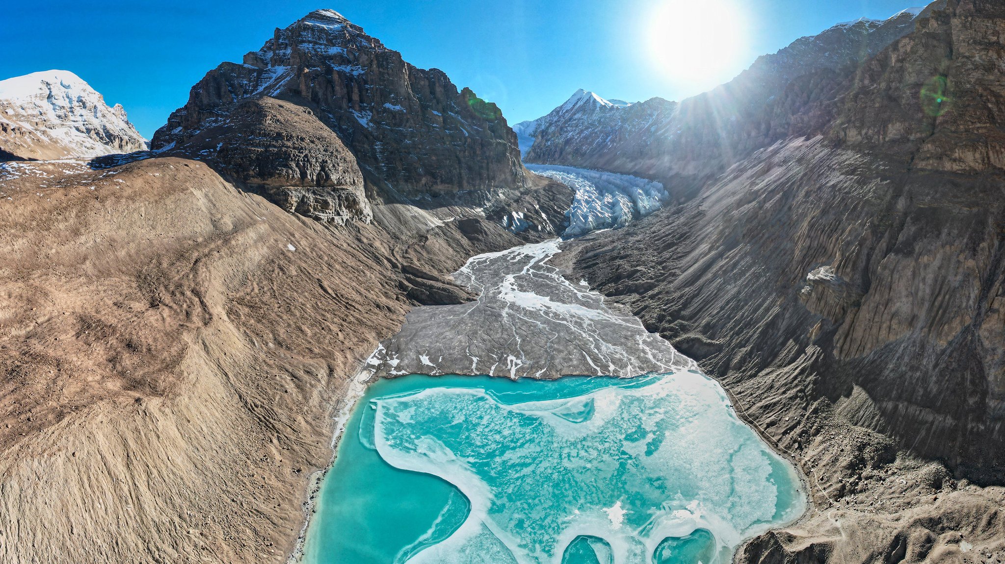 Gletscherzunge mit braunem Gebirge, blauer Himmel, Sonne; die Landschaft des Qoidenyima-Gletschers im Kreis Gangba der Stadt Xigaze in der autonomen Region Tibet im Südwesten Chinas.