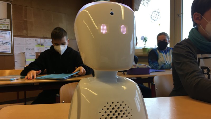 Ein weißer Roboter steht auf einer Schulbank in einem Klassenzimmer.