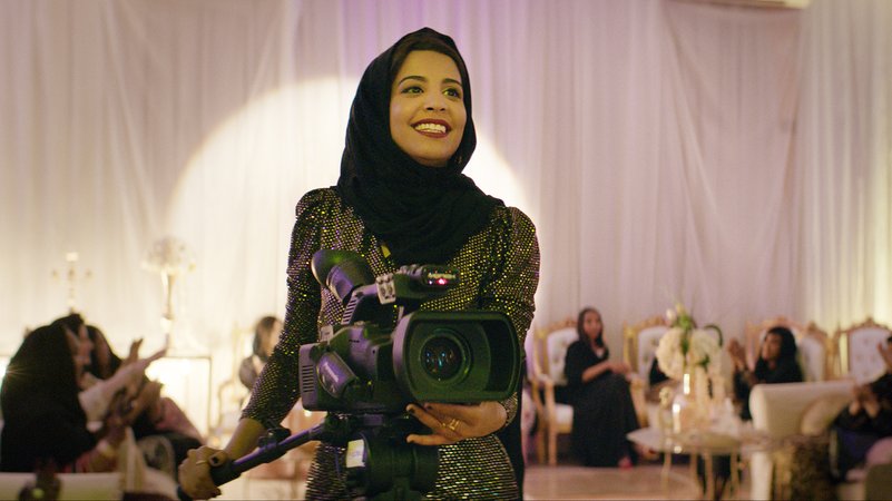 Eine Frau in einem Glitzer-Kleid und mit schwarzem Kopftuch lächelt und bedient eine Filmkamera: Szene aus "Die perfekte Kandidatin"